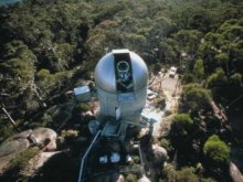 Birds eye view of the Orroral SLR Observatory. It is a dome shaped building approximately 15 m wide with a telescope visible through the top. The Observatory is built on rock in a clearing within bushland.